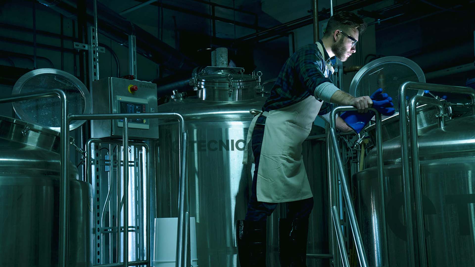 A biotechnologist wearing a white lab coat and safety glasses, standing beside a large stainless steel fermentation tank in an industrial facility