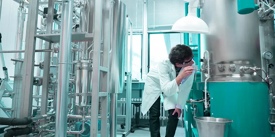 A person working next to a stainless steel fermenter in a room with other biotechnological equipment, cover of difference between a fermenter and a bioreactor blog